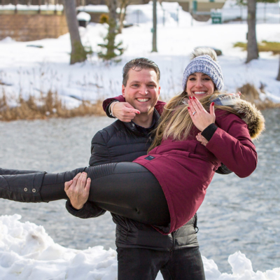 Man holding up his fiance, who is pointing to an engagement ring.