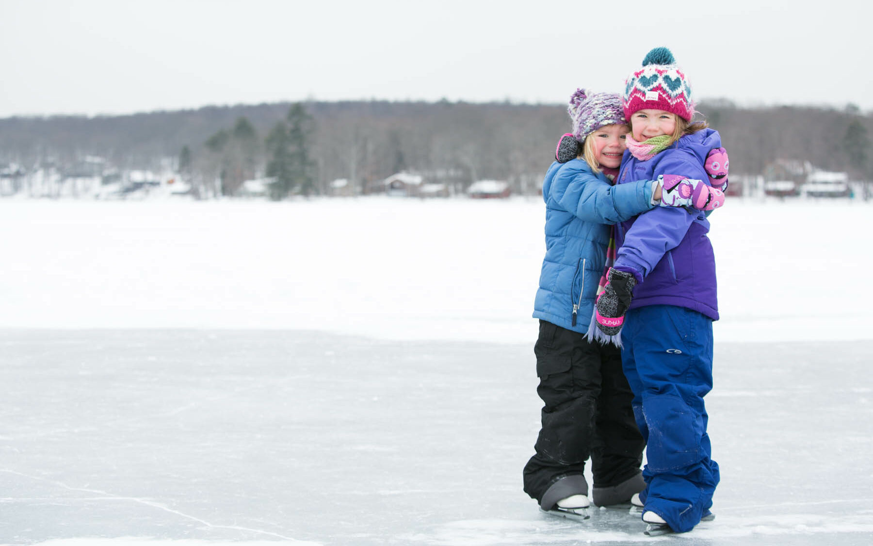 Two young girls in snow suits and ice skates hugging on Lake Teedyuskung in winter.